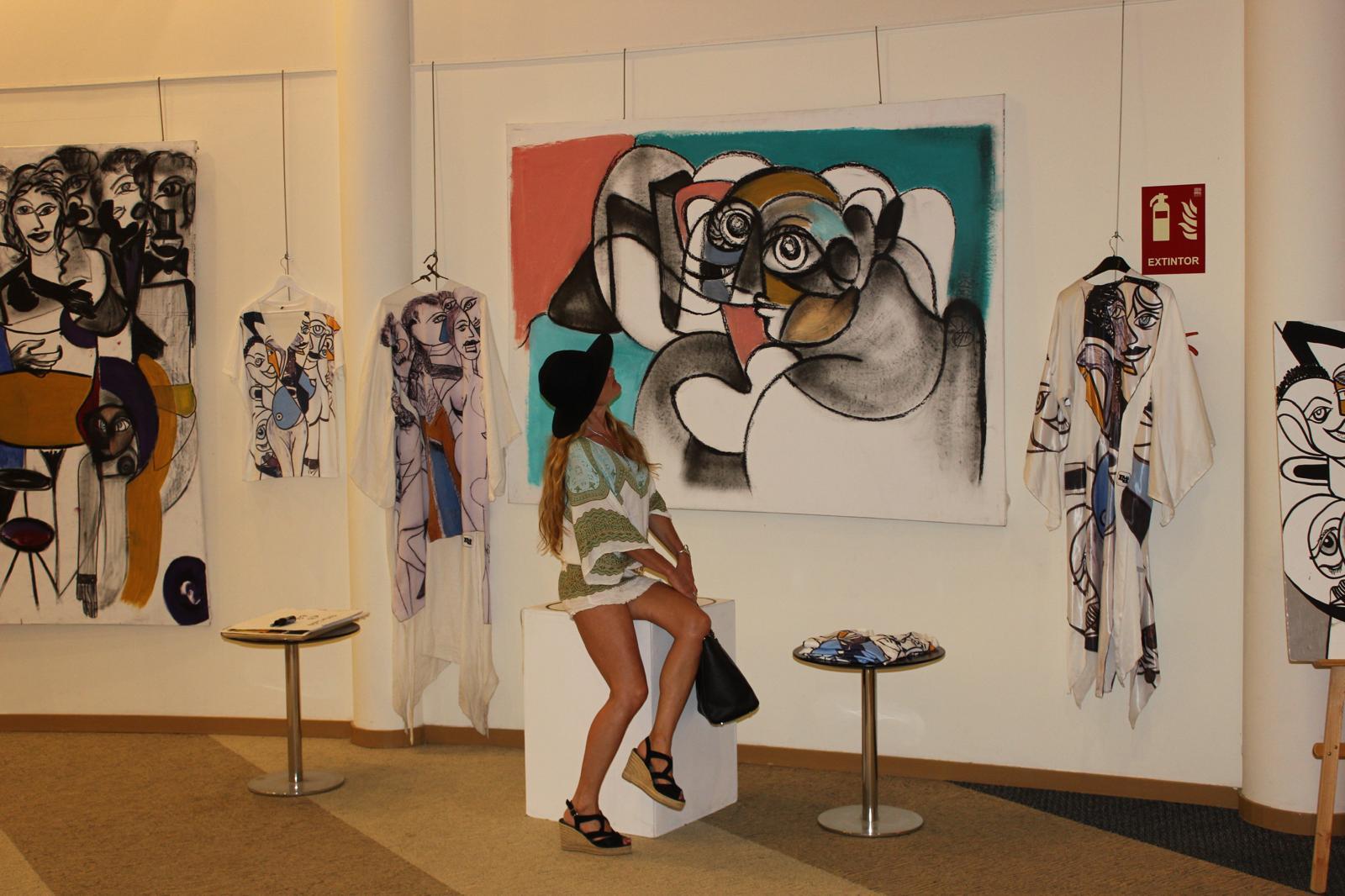 Painted garments hung beside the canvases at an exhibition, a visitor seated between them