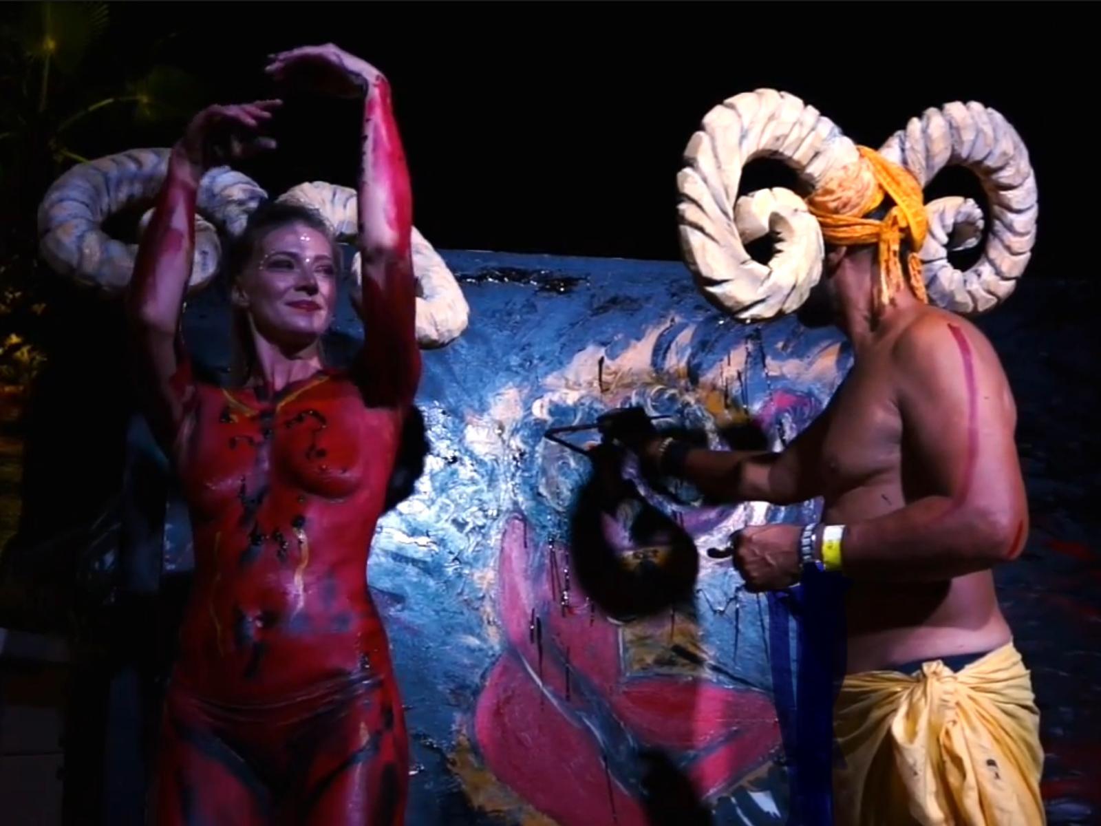 Two body-painted performers in horned headdresses beside a sculptural canvas at night