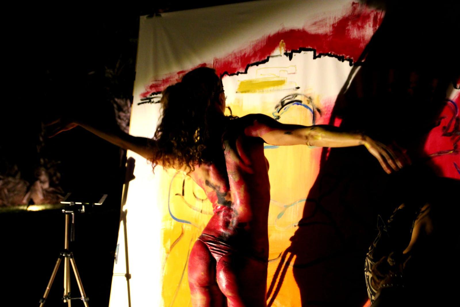 A body-painted performer before a red and yellow canvas, lit against the dark